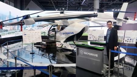 　スカイドライブがジャパンモビリティショーで展示する、空飛ぶクルマの模型と自動改札機。右は福沢知浩ＣＥＯ＝２９日午前、東京都江東区の東京ビッグサイト