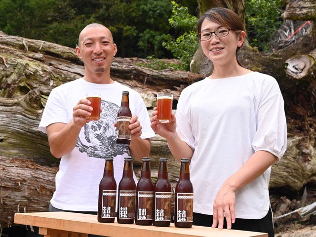 倒れた大杉の前で完成したビールを手にする田村啓さんと東恵理子社長（右）＝瑞浪市大湫町、大湫神明神社