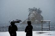 　雪が降りしきる中、石川県珠洲市の景勝地「見附島」を訪れた人たち。能登半島地震の発生から２年となった＝１日午前７時５分