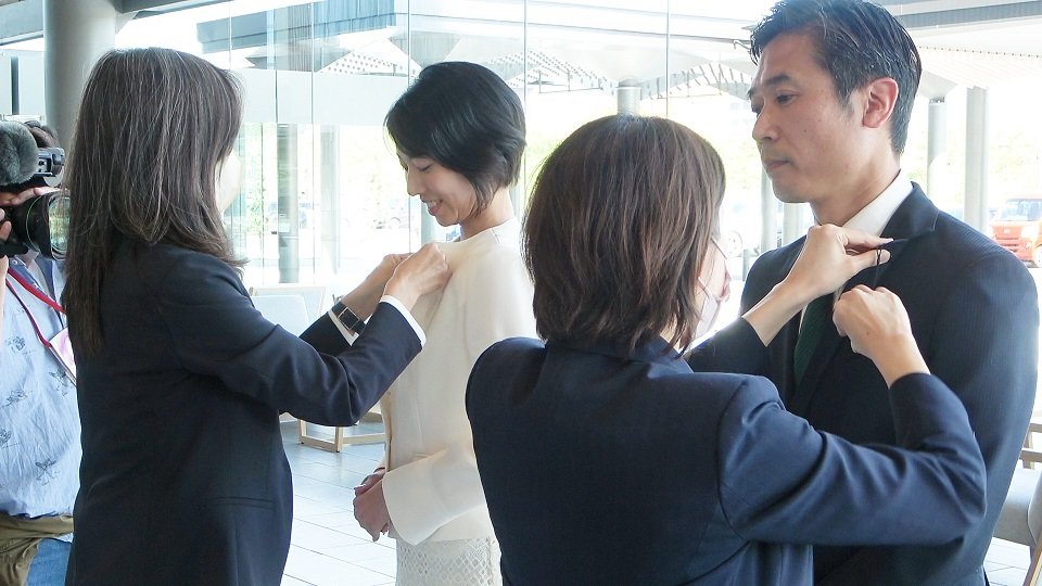 襟にバッジを着けてもらう判治康信議員（右）と今井瑠々議員＝県議会棟