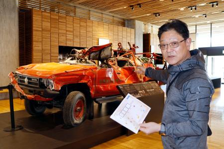 　東日本大震災津波伝承館の被災消防車の前で、謎解きについて説明する「キャピタルホテル１０００」の松田修一社長＝４月、岩手県陸前高田市