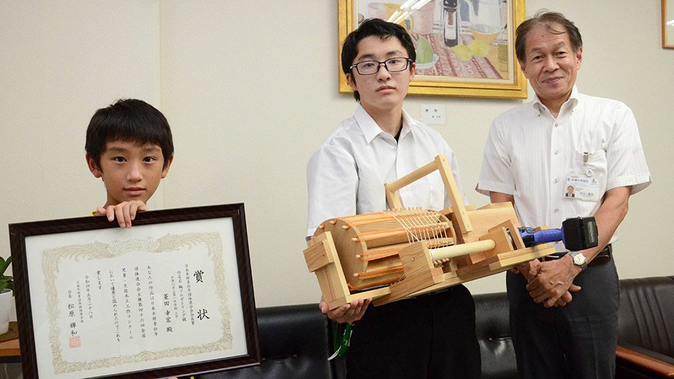 入賞作品の「輪ゴムガトリング銃」を抱え、製作の苦労を振り返った菱田幸宏さん（中央）＝中津川市栄町、市にぎわいプラザ
