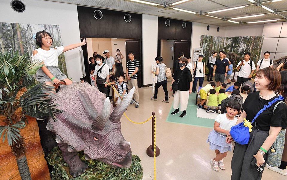 トリケラトプスのロボットの背に乗り、笑顔を見せる女の子＝岐阜市本荘、市科学館（撮影・山田雄大）