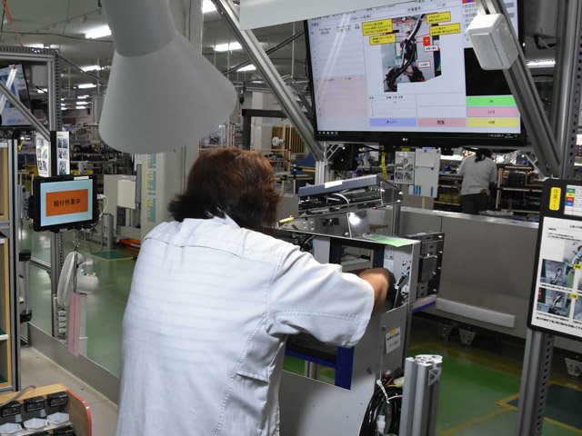 頭上のモニターに表示された手順に従い、運賃箱を組み立てる従業員